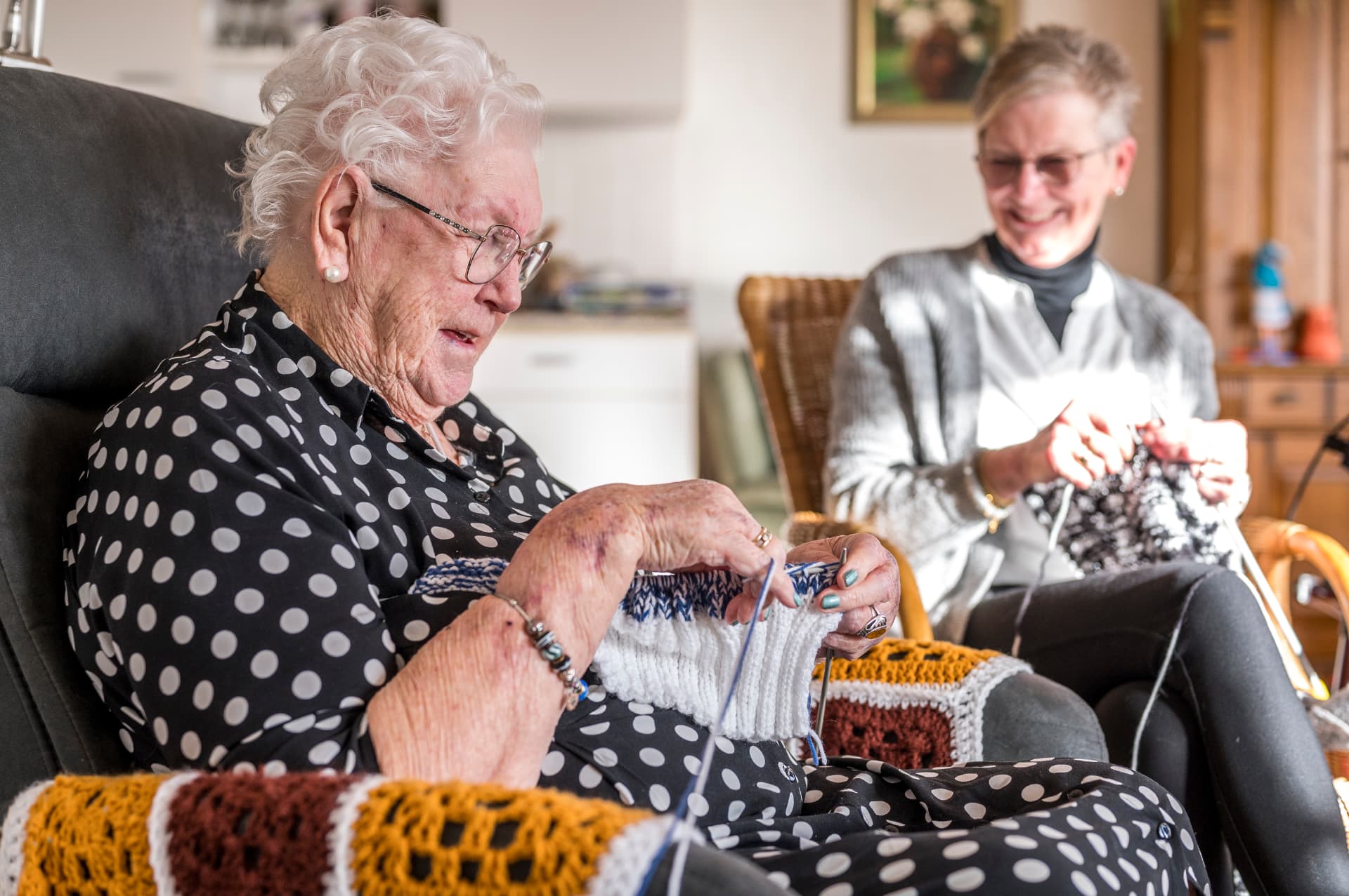 Oudere vrouw zit te breien in haar stoel, met bezoek dat naast haar meebreit