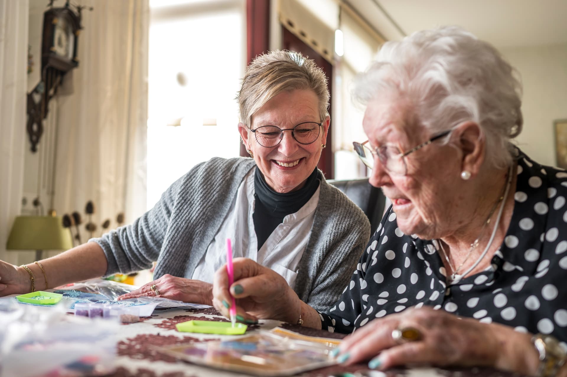 Een vrijwilliger en een oudere vrouw maken samen lachend een diamond painting aan de tafel
